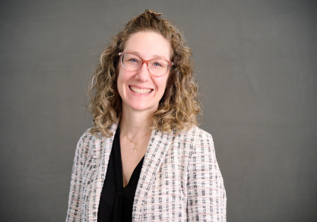 Woman with curly hair and glasses, wearing a light-colored checkered blazer over a black top, smiling in front of a plain gray background